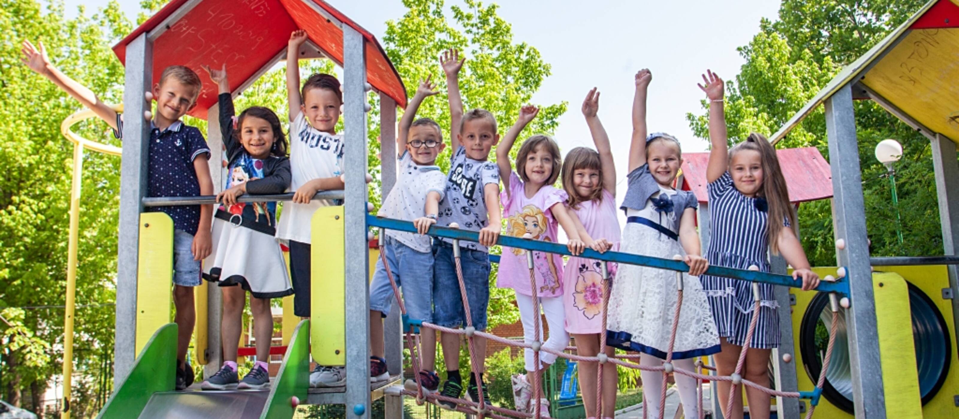 Neun Kinder winken lachend von einem Rutschbahnturm auf einem Spielplatz.